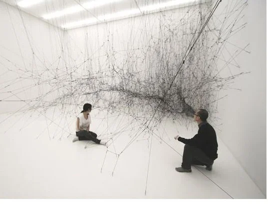 Two people sit opposite each other with a web of threads stretched between them.